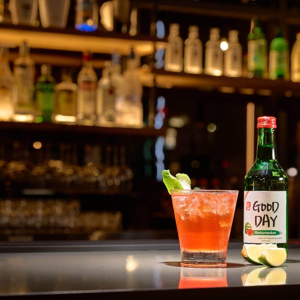 Watermelon soju cocktail with lime garnish and a bottle of Good Day Watermelon Soju on a bar.