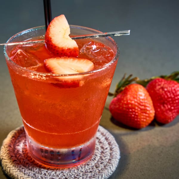 A vibrant red strawberry cocktail with ice and fresh strawberry slices, on a woven coaster.