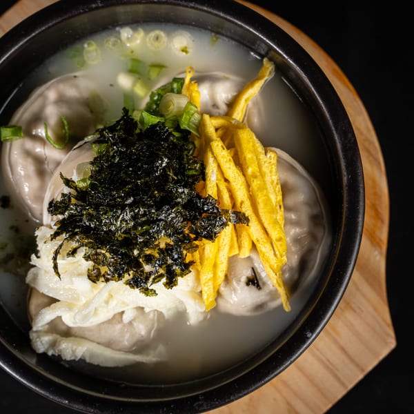 Korean Mandu-guk (dumpling soup) with seaweed, egg strips, and green onions in a black bowl.