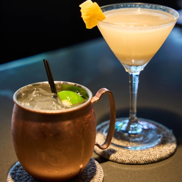 Two cocktails on a dark bar: a Moscow Mule and a martini with a lemon peel garnish.