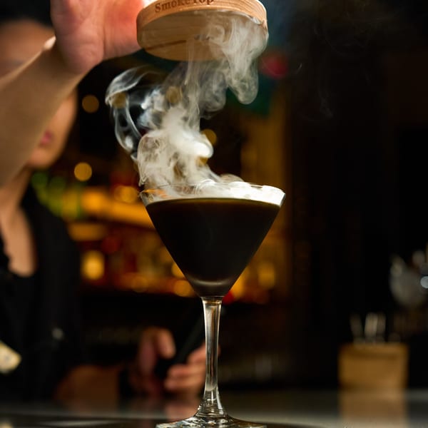 A dark cocktail in a martini glass being smoked by a wooden lid held by a hand.