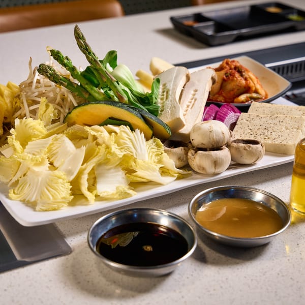 A white platter of fresh vegetables, tofu, and kimchi for Korean BBQ, with dipping sauces.