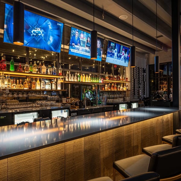 A well-stocked bar at SSAMJANG Korean BBQ with multiple TVs showing sports and news, and bar stools.